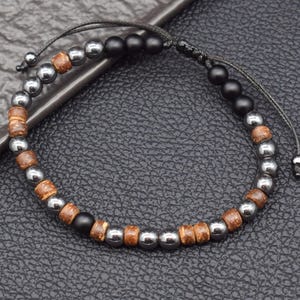 Könnte beinhalten: Verstellbares Armband mit einer Mischung aus Perlen. Das Armband besteht aus silberfarbenen, schwarzen und braunen Holzperlen. Die Perlen sind auf einer schwarzen Kordel aufgefädelt, mit verstellbaren Gleitknoten für eine individuelle Passform. Das Armband ist für den täglichen Gebrauch konzipiert.