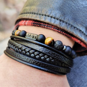 May include: A layered bracelet set with black leather bands, black and brown beads, and a silver charm engraved with "M&T".