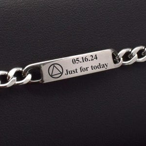 Puede incluir: Una pulsera de cadena de plata con una placa rectangular grabada. La placa tiene grabado el texto "05.16.24", un triángulo dentro de un círculo y "Just for today". La pulsera se muestra sobre una superficie negra.