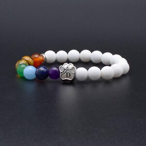 May include: A stretch bracelet featuring matte white beads and a series of colorful gemstone beads including purple, blue, green, yellow, and orange. A silver-toned metal charm shaped like a paw print with the letter D engraved in the center.