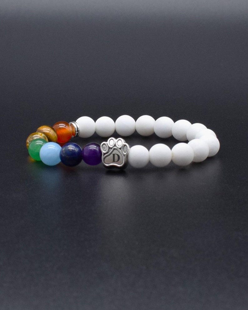 May include: A stretch bracelet featuring matte white beads and a series of colorful gemstone beads including purple, blue, green, yellow, and orange. A silver-toned metal charm shaped like a paw print with the letter D engraved in the center.
