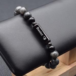 May include: A black beaded bracelet with a silver charm engraved with the name "Buster" and a paw print. The bracelet is on a black display stand.