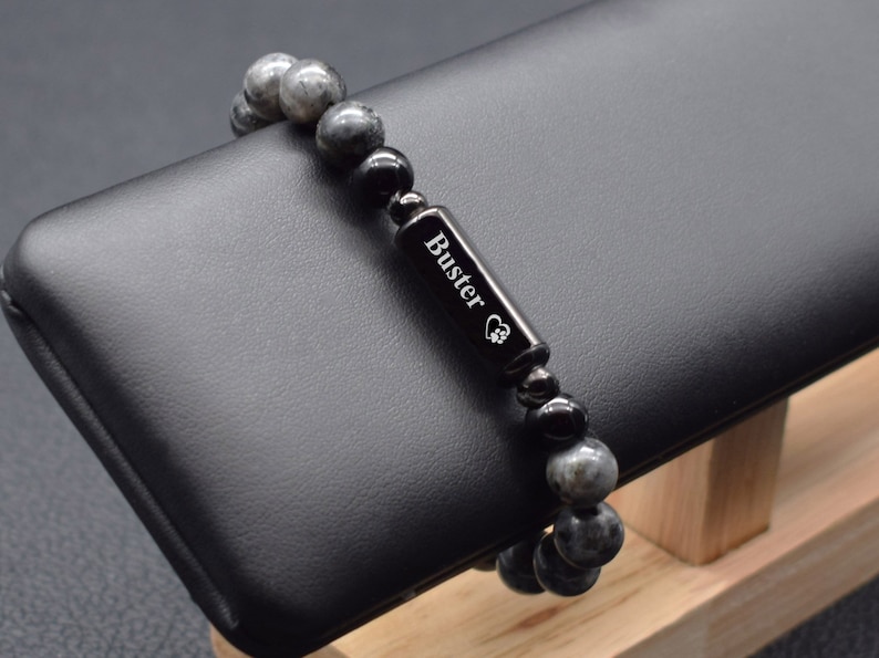 May include: A black beaded bracelet with a silver charm engraved with the name "Buster" and a paw print. The bracelet is on a black display stand.