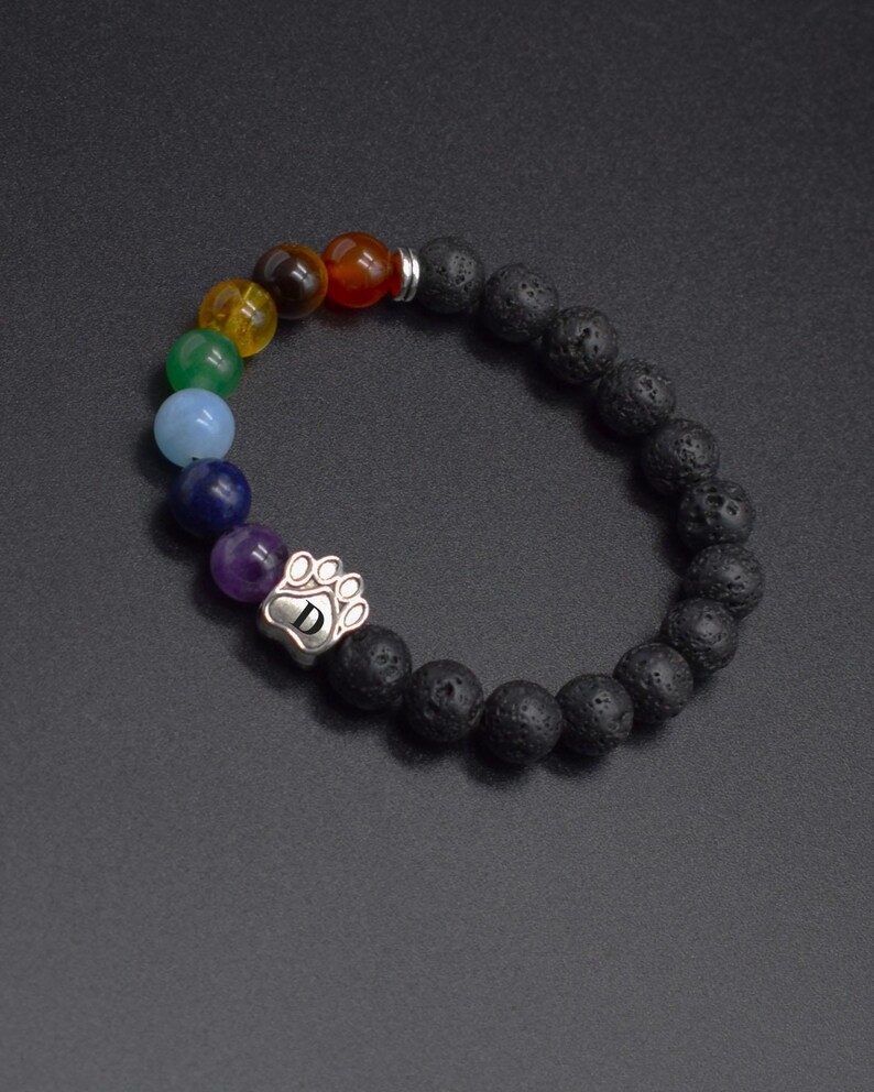 May include: A beaded bracelet featuring black porous lava stone beads and a series of smooth, multi-colored gemstone beads. A silver-toned metal charm shaped like a paw print with the letter D engraved in the center is integrated into the design.