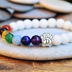 May include: A beaded bracelet featuring matte white beads and a sequence of colorful accent beads including red, orange, yellow, green, light blue, dark blue, and purple. A silver-toned metal charm shaped like a paw print with the letter D is included.