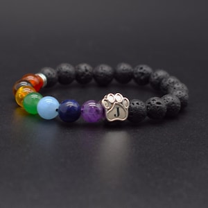May include: A stretch bracelet featuring black porous lava stone beads, a silver-toned metal paw print charm engraved with the letter J, and a sequence of smooth, multi-colored gemstone beads in red, orange, yellow, green, light blue, dark blue, and purple.