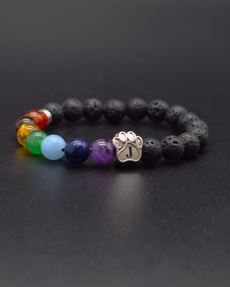 May include: A stretch bracelet featuring black porous lava stone beads, a silver-toned metal paw print charm engraved with the letter J, and a sequence of smooth, multi-colored gemstone beads in red, orange, yellow, green, light blue, dark blue, and purple.