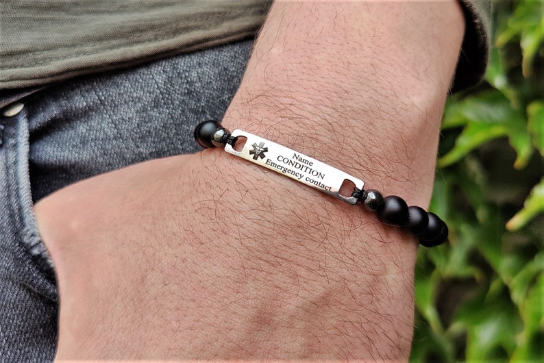 May include: A medical alert bracelet featuring matte black beads and a silver-toned metal rectangular plate. The plate displays a medical star of life symbol and the engraved text: Name, CONDITION, Emergency contact.