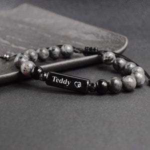 May include: A black beaded bracelet with a black rectangular charm engraved with the name "Teddy" and a paw print inside a heart. The bracelet is made with grey and black beads.