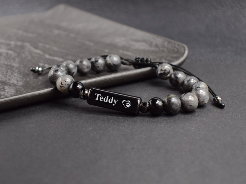 May include: A black beaded bracelet with a black rectangular charm engraved with the name "Teddy" and a paw print inside a heart. The bracelet is made with grey and black beads.