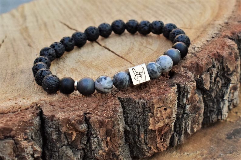 May include: Black lava rock and grey agate beaded bracelet with a silver charm featuring a rock and roll hand gesture.