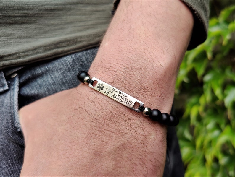 May include: A medical alert bracelet featuring matte black beads and a rectangular silver-toned metal plate. The plate displays a medical star of life symbol and the engraved text: Jack White, TYPE 1 DIABETIC, ICE: 786-875-9374.
