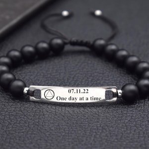 Può includere: Braccialetto con perline nere e un ciondolo argentato con la scritta "07.11.22 One day at a time" e un simbolo triangolare.