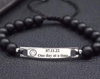 May include: A beaded bracelet featuring matte black spherical beads and a central silver-toned metal bar. The bar is engraved with a triangle symbol, the date 07.11.22, and the text One day at a time. The bracelet uses a black cord closure.