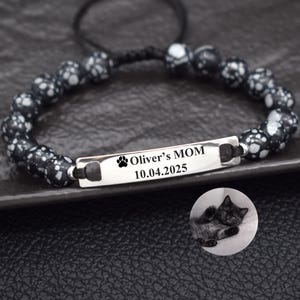 May include: A black and white beaded bracelet with a silver bar engraved with "Oliver's MOM", a paw print, and the date 10.04.2025. A small circular image of a black cat is in the background.