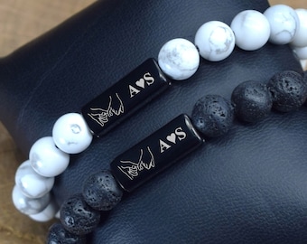 May include: Two bracelets with black lava rock and white howlite beads. One bracelet has a black rectangular charm with the inscription "A♥S" and a hand-holding design. The other bracelet has a similar charm with the inscription "A♥S" and a hand-holding design.