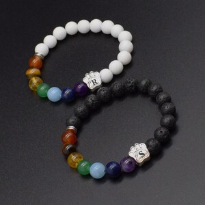 May include: Two beaded bracelets featuring a row of colorful chakra stones and a silver-toned paw print charm. One bracelet has matte white beads, and the other has textured black lava beads. The paw charms are engraved with the letters R and S.