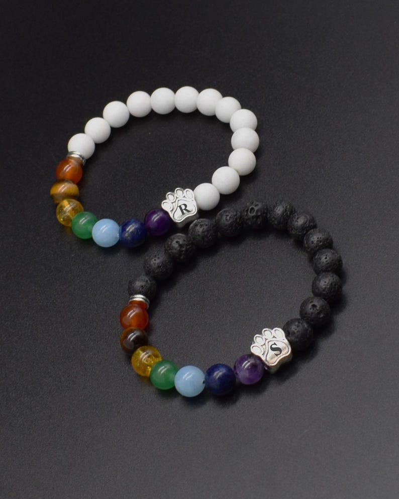May include: Two beaded bracelets featuring a row of colorful chakra stones and a silver-toned paw print charm. One bracelet has matte white beads, and the other has textured black lava beads. The paw charms are engraved with the letters R and S.