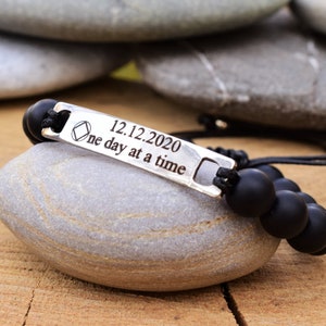 May include: A black beaded bracelet featuring a silver-toned metal bar engraved with a diamond-shaped symbol, the date 12.12.2020, and the text "one day at a time". The bracelet is constructed with matte black spherical beads and black cord.
