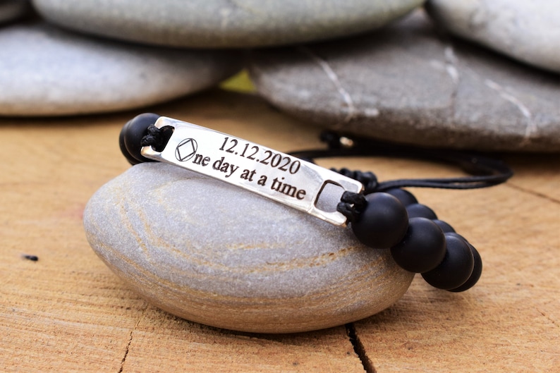 May include: A black beaded bracelet featuring a silver-toned metal bar engraved with a diamond-shaped symbol, the date 12.12.2020, and the text "one day at a time". The bracelet is constructed with matte black spherical beads and black cord.