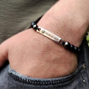 May include: A medical alert bracelet featuring a rectangular silver-toned metal plate engraved with the Star of Life symbol and the text Medical alert. The plate is connected to a strand of matte black beads and two polished dark grey beads.
