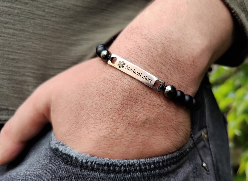 May include: A medical alert bracelet featuring a rectangular silver-toned metal plate engraved with the Star of Life symbol and the text Medical alert. The plate is connected to a strand of matte black beads and two polished dark grey beads.
