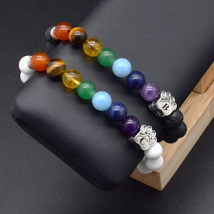 May include: Two beaded bracelets featuring a sequence of colorful gemstone beads in red, brown, yellow, green, light blue, dark blue, and purple. Each bracelet includes a silver-toned metal charm shaped like a paw print with the letters B or C.