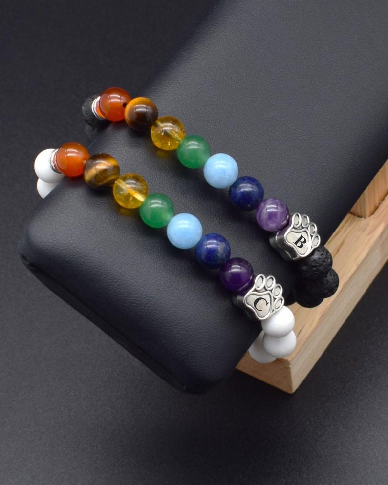 May include: Two beaded bracelets featuring a sequence of colorful gemstone beads in red, brown, yellow, green, light blue, dark blue, and purple. Each bracelet includes a silver-toned metal charm shaped like a paw print with the letters B or C.