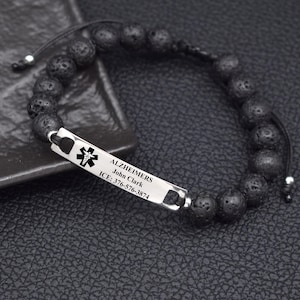 May include: A medical alert bracelet featuring black porous lava stone beads on a black cord. A rectangular silver-tone metal plate is engraved with a black medical caduceus symbol, the text ALZHEIMERS, John Clark, and ICE: 376-576-3874.
