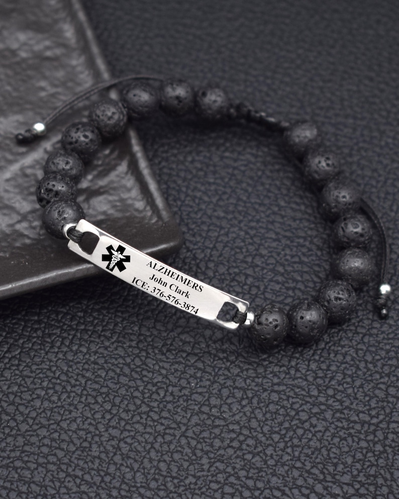 May include: A medical alert bracelet featuring black porous lava stone beads on a black cord. A rectangular silver-tone metal plate is engraved with a black medical caduceus symbol, the text ALZHEIMERS, John Clark, and ICE: 376-576-3874.