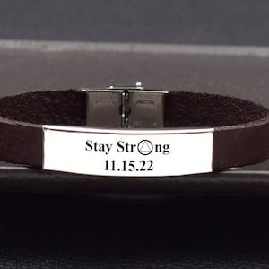 May include: Brown leather bracelet with a silver plate that reads "Stay Strong 11.15.22" and a triangle symbol.