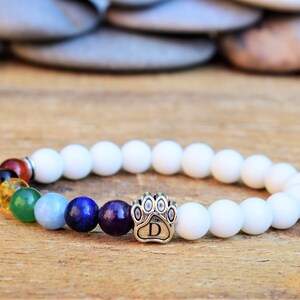 May include: A stretch bracelet featuring matte white beads and a series of seven colorful gemstone beads in red, black, yellow, green, light blue, dark blue, and purple. A silver-toned metal paw print charm with the letter D is centered.