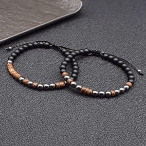 May include: Two beaded bracelets. Each bracelet features black matte beads, interspersed with silver-colored metal beads and sections of brown wooden beads. The bracelets are on a black surface.