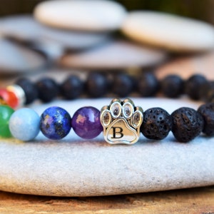 May include: A beaded bracelet featuring black porous lava stone beads, a series of smooth multi-colored chakra beads, and a silver-toned metal charm shaped like a paw print with the letter B engraved in the center of the paw pad.