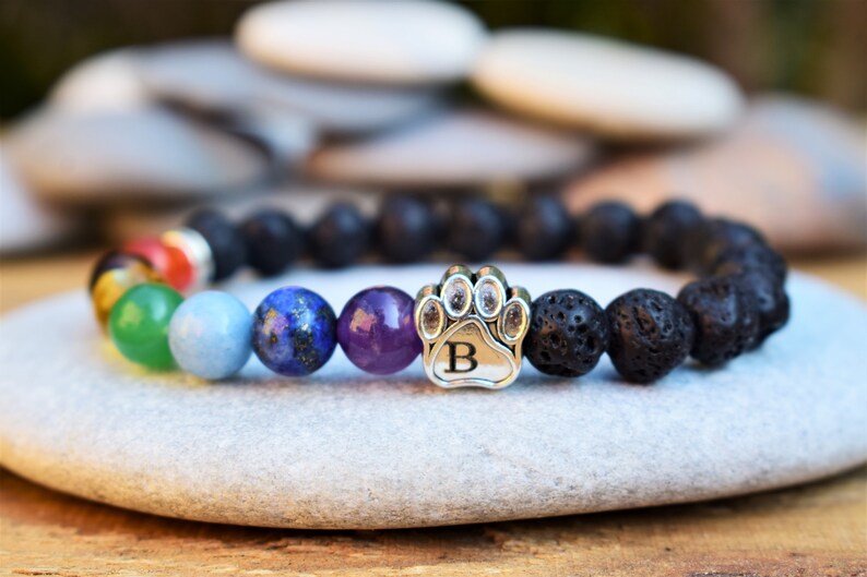 May include: A beaded bracelet featuring black porous lava stone beads, a series of smooth multi-colored chakra beads, and a silver-toned metal charm shaped like a paw print with the letter B engraved in the center of the paw pad.