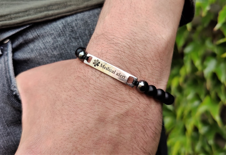May include: A medical alert bracelet featuring a rectangular silver-toned metal plate engraved with the text Medical alert and a star of life symbol. The plate is connected to a strand of smooth black beads and dark metallic spherical beads.