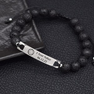 May include: A black beaded bracelet featuring a rectangular silver-toned metal bar engraved with a triangle inside a circle symbol and the text 1 Year Sober 06.12.22. The bracelet is constructed with porous black beads and a black cord closure.