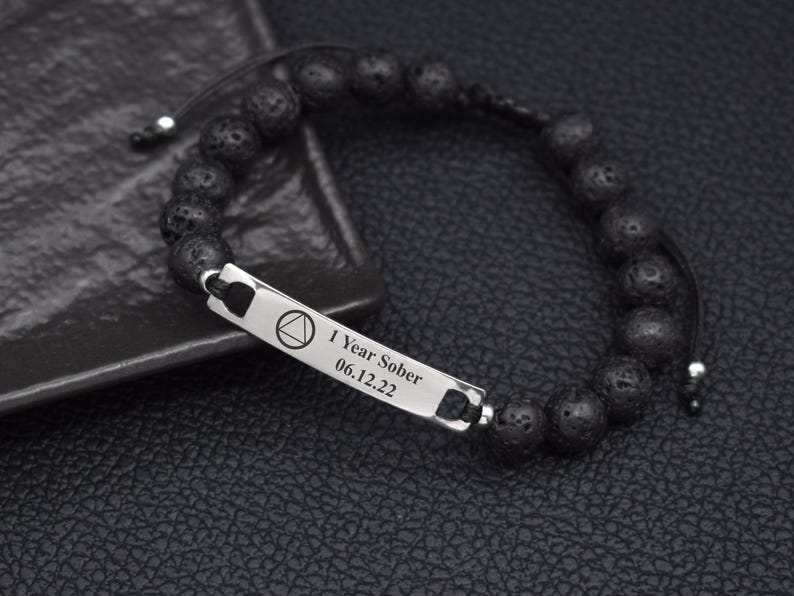 May include: A black beaded bracelet featuring a rectangular silver-toned metal bar engraved with a triangle inside a circle symbol and the text 1 Year Sober 06.12.22. The bracelet is constructed with porous black beads and a black cord closure.