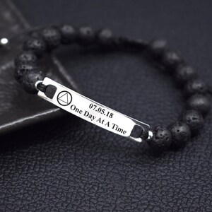 May include: A black beaded bracelet featuring a rectangular silver-toned metal bar. The bar is engraved with a triangle symbol, the date 07.05.18, and the text One Day At A Time. The beads have a porous, textured surface and are strung on a cord.