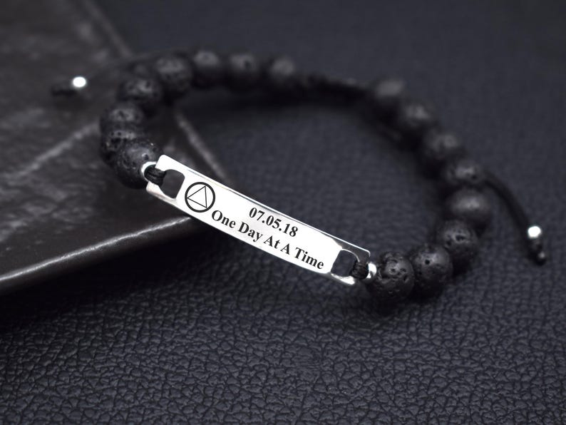 May include: A black beaded bracelet featuring a rectangular silver-toned metal bar. The bar is engraved with a triangle symbol, the date 07.05.18, and the text One Day At A Time. The beads have a porous, textured surface and are strung on a cord.