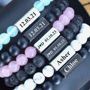 May include: Five bracelets with different colored beads and silver metal tags. The tags have the dates "12.03.21", "12.03.21", "JVD 01.10.23", "JVD 01.10.23", and the names "Asher" and "Chloe".