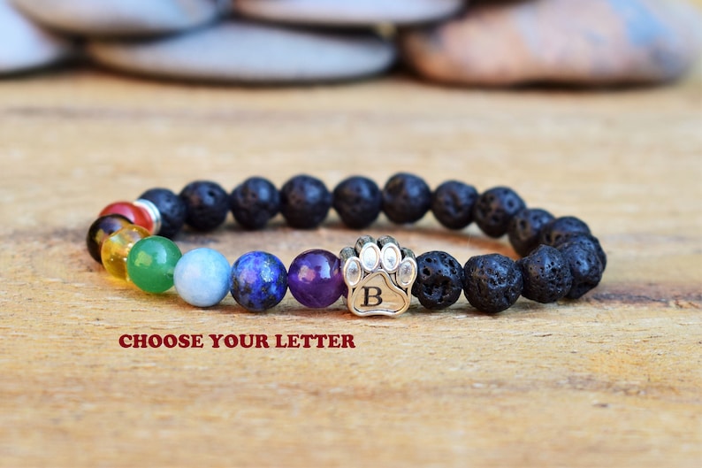 May include: A beaded bracelet featuring black porous lava stone beads, a series of smooth multi-colored chakra beads, and a silver-toned metal paw print charm with the letter B engraved in the center. The text CHOOSE YOUR LETTER is visible.