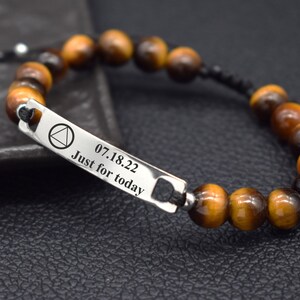 May include: A beaded bracelet featuring brown and black tiger eye stones strung on a black cord. A rectangular silver-toned metal bar is integrated into the design, engraved with a triangle symbol, the date 07.18.22, and the text Just for today.