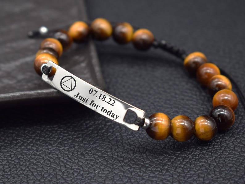 May include: A beaded bracelet featuring brown and black tiger eye stones strung on a black cord. A rectangular silver-toned metal bar is integrated into the design, engraved with a triangle symbol, the date 07.18.22, and the text Just for today.