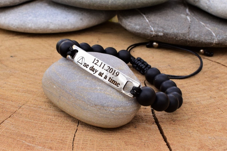 May include: A black beaded bracelet featuring a silver-toned metal bar engraved with a triangle symbol, the date 12.11.2019, and the text "one day at a time". The bracelet is constructed with matte black round beads and a black cord closure.