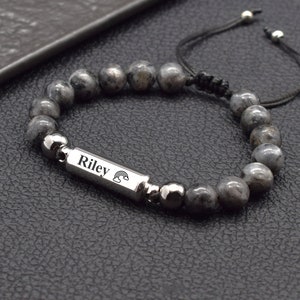 May include: A grey beaded bracelet with a silver charm that says "Riley" with a rainbow design. The bracelet is made with a black cord and silver beads.