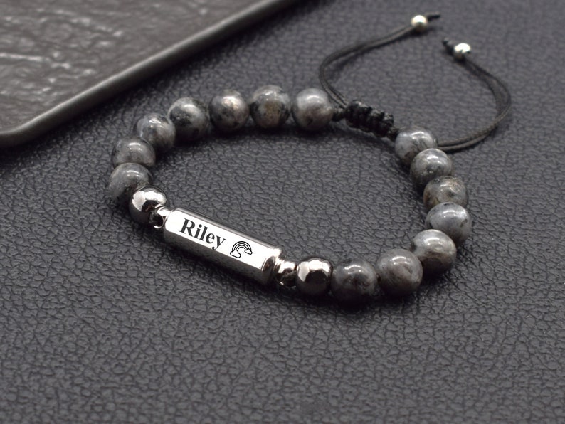May include: A grey beaded bracelet with a silver charm that says "Riley" with a rainbow design. The bracelet is made with a black cord and silver beads.