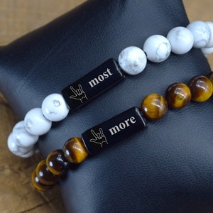 May include: Two bracelets with black metal tags that say "most" and "more" in white lettering. The bracelet on the left is made of white beads and the bracelet on the right is made of brown tiger's eye beads.
