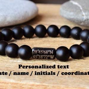 Puede incluir: Una pulsera elástica compuesta por cuentas esféricas de color negro mate. Una cuenta rectangular central de color negro presenta el texto blanco grabado 28°45'68"N 45°38'72"W. Debajo de la pulsera, el texto dice: Personalized text, date / name / initials / coordinates.