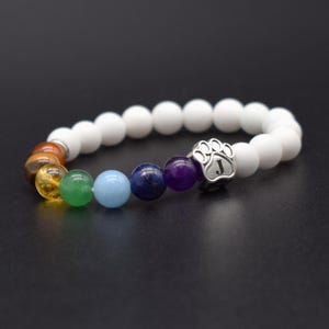 May include: A beaded stretch bracelet featuring matte white beads and a series of colorful gemstone beads in shades of orange, yellow, green, light blue, dark blue, and purple. A silver-toned metal charm shaped like a paw print with the letter J.